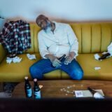 Un bărbat obosit, așezat pe o canapea seara, cu partenera care îi pune o mână pe umăr în semn de sprijin