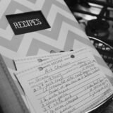Caiet vechi cu rețete scrise de mână, deschis pe o masă de bucătărie lângă făină, ouă și un făcăleț