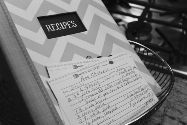 Caiet vechi cu rețete scrise de mână, deschis pe o masă de bucătărie lângă făină, ouă și un făcăleț