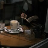 O persoană care citește liniștită la o masă de cafenea, lângă o fereastră luminoasă, așteptând o întâlnire