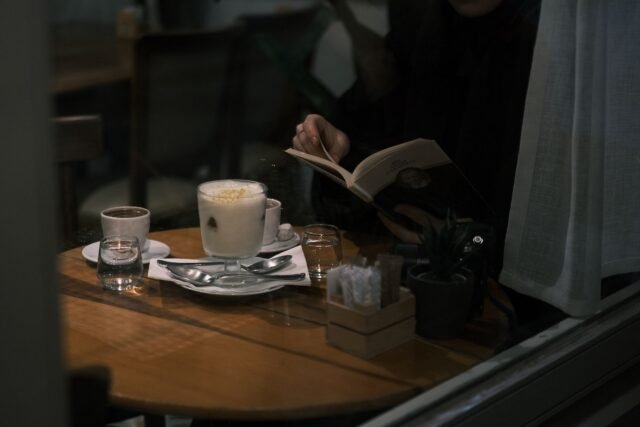 O persoană care citește liniștită la o masă de cafenea, lângă o fereastră luminoasă, așteptând o întâlnire