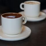 Două cești de cafea pe o măsuță de cafenea, lângă fereastră, pregătite pentru o primă întâlnire
